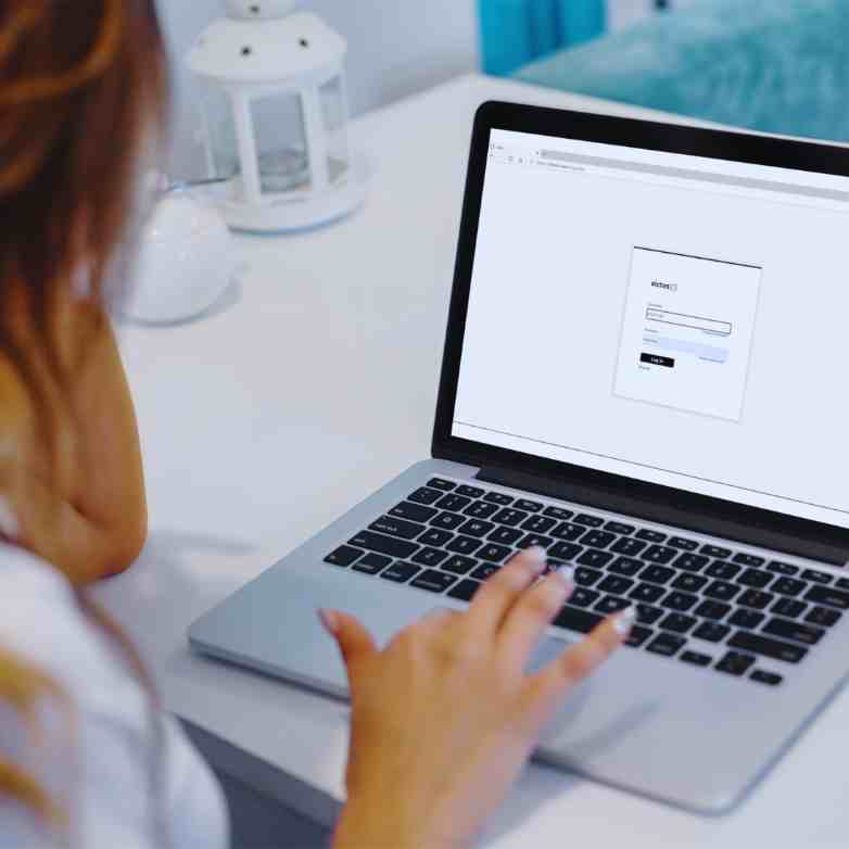 White woman at a laptop, facing away from the camera and looking at a screen with a login box open.