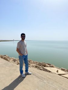 A Middle Eastern man stands in front of the sea, wearing blue jeans, a grey shirt and sunglasses.